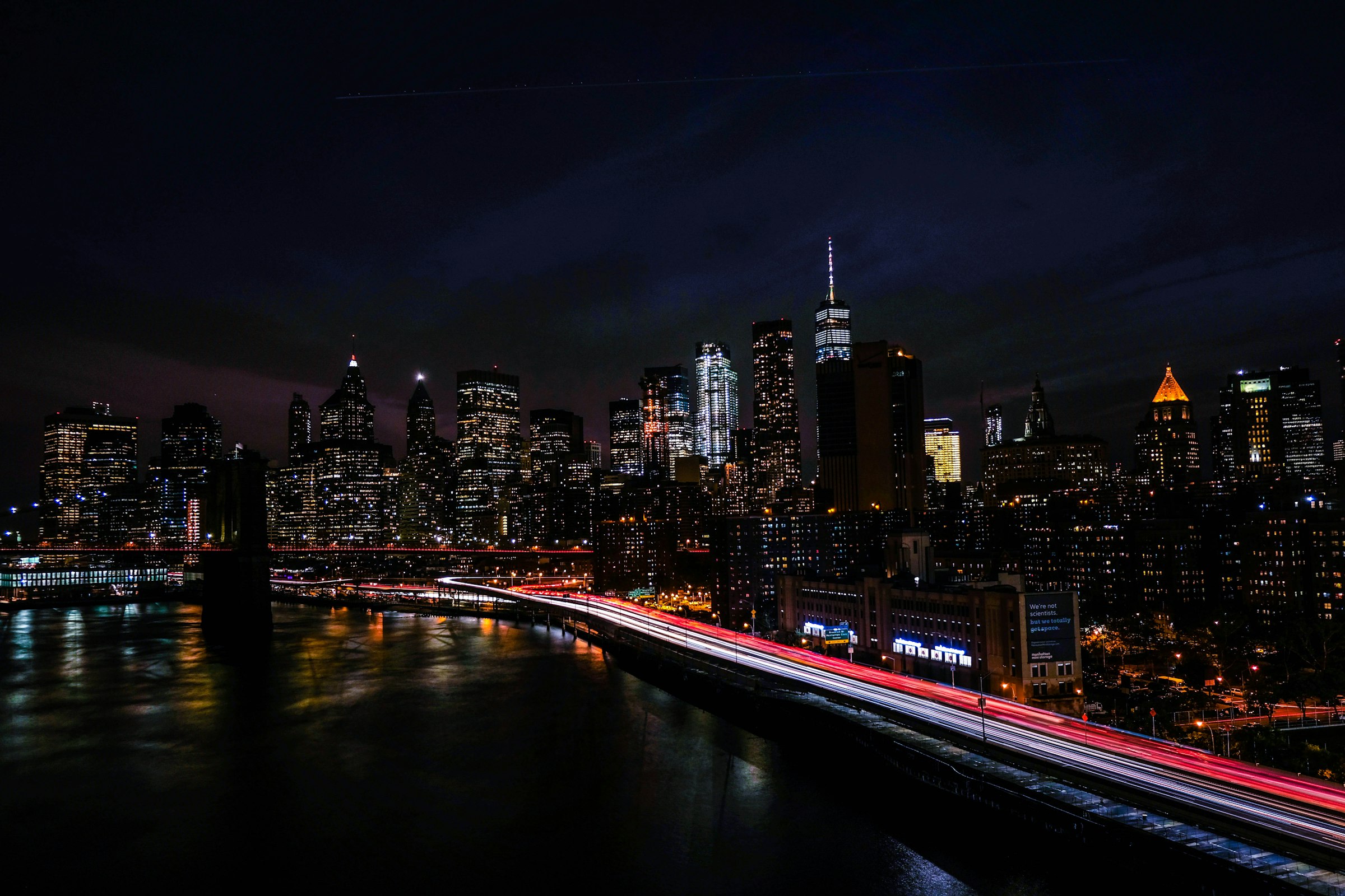 The Lower Manhattan skyline at night, with light trails along the riverside highway